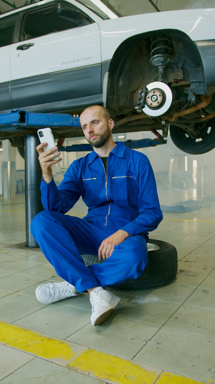 services-04 Mechanic in a blue jumpsuit sitting on a tire, using a smartphone, with a car in repair overhead.